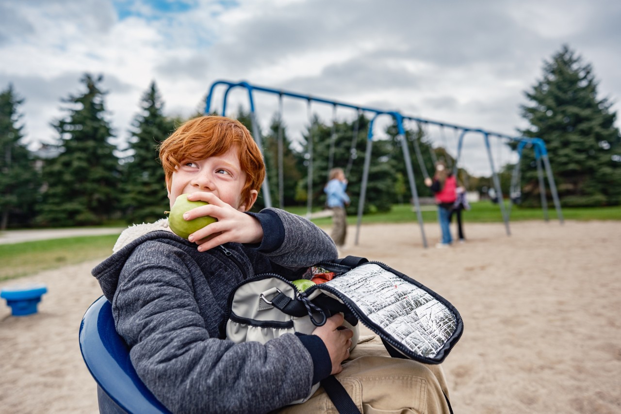 School age boy having a fruit snack in the park. Girls on the swing in the background. Exterior of public park in Mississauga, Ontario.
