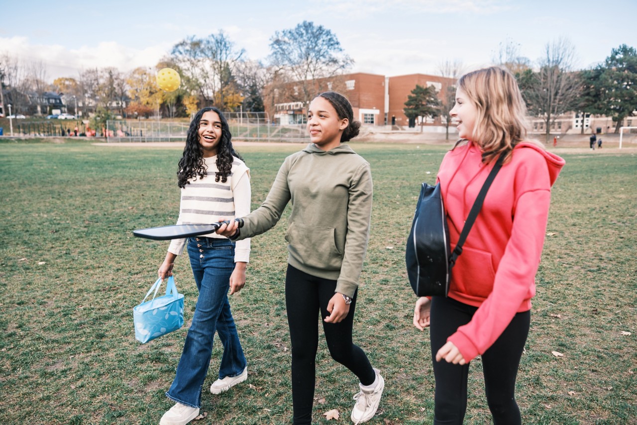 Multiracial group of school age girls on the way to play pickle-ball outdoors in urban city park. All dressed in casual clothes.